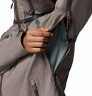 A person wearing a taupe Columbia Women’s Reign No Shine Waterproof Jacket unzips a large side zipper, revealing light green inner lining and ventilation mesh; the image highlights hand and jacket details against a plain backdrop.