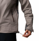 A person tightens the drawcord at the hem of a taupe Columbia Womens Reign No Shine Waterproof Jacket for better rain protection. Their torso and hand are shown against a white background.