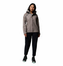 A woman faces forward against a white background, wearing the Columbia Womens Reign No Shine Waterproof Jacket in gray, paired with black pants and black sneakers with light green soles, ready for rain protection.