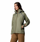 A woman in a Columbia Womens Reign No Shine Waterproof Jacket in light olive green with zippered pockets and tan pants stands angled against a plain white background, ready for rain protection.