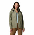 A woman with long brown hair models the Columbia Womens Reign No Shine Waterproof Jacket in light olive green over a light blue top and brown pants, standing against a plain white background.
