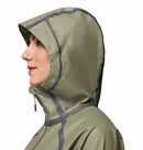 A woman in profile wears a light olive green Columbia Womens Reign No Shine Waterproof Jacket with dark gray sealed seams and zipper, hood up for maximum rain protection.