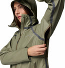 A person wearing a Columbia Womens Reign No Shine Waterproof Jacket in olive unzips an underarm vent. The rain-ready jacket has dark waterproof seams, multiple zippers, and the persons partially visible face looks to the left.
