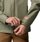 A woman adjusts the cuff of a light olive green Columbia Womens Reign No Shine Waterproof Jacket with a velcro strap. The jacket features a red and white Outdoor patch on the sleeve, paired with light brown pants for added rain protection.
