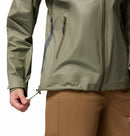 A person adjusts the hem of a Columbia Womens Reign No Shine Waterproof Jacket, shown in green, highlighting its rain protection. Only the torso and part of the left arm are visible above brown pants.