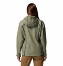 A woman with long brown hair, wearing the Columbia Womens Reign No Shine Waterproof Jacket in olive green and tan pants, stands facing away from the camera against a plain white background.
