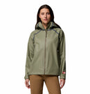 A woman stands facing forward against a plain white background, wearing the Columbia Womens Reign No Shine Waterproof Jacket in light olive green with a hood and front zipper, paired with brown pants.