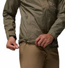 A man in tan pants tightens the waist drawstring of a Columbia Mens Pouring Adventure™ III Waterproof Hiking Jacket, shown in light olive green with focus on the hands and lower half, highlighting its versatility for outdoor use.