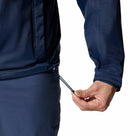 A hand tightens the drawstring at the hem of a Columbia Mens Pouring Adventure™ III Waterproof Hiking Jacket in dark blue. Only the lower part of this packable rain jacket and the person’s hand are visible.