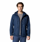 A young man stands facing forward, wearing the Columbia Mens Pouring Adventure™ III Waterproof Hiking Jacket in navy blue over a gray t-shirt and blue pants against a plain white background.