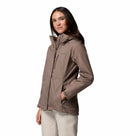 A woman with long brown hair, dressed in light pants, stands against a white background wearing a taupe Columbia Womens Pouring Adventure III Waterproof Hiking Jacket with her hands in the pockets.