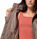 A woman opens her taupe Columbia Womens Pouring Adventure III Waterproof Hiking Jacket, revealing its mesh lining. She wears a sleeveless coral top, and the cropped image shows her from shoulders to mid-torso.