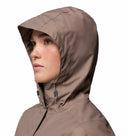 A woman is shown in profile wearing the Columbia Womens Pouring Adventure III Waterproof Hiking Jacket in light brown. The hood is up, covering most of her head, with adjustable drawstrings at the neck. The background is plain white.