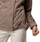 A person, seen from the waist down, adjusts the hem drawstring on a Columbia Womens Pouring Adventure III Waterproof Hiking Jacket in brown, paired with light pants. Only their lower torso and one hand are visible.