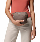 A person in a sleeveless coral top and beige pants holds a compact brown mesh travel bag with a visible zipper and brand label, ideal for carrying essentials like the Columbia Womens Pouring Adventure III Waterproof Hiking Jacket.