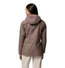 A woman with long brown hair stands with her back to the camera against a white background, wearing light-colored pants and a taupe Columbia Womens Pouring Adventure III Waterproof Hiking Jacket.