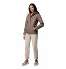 A woman stands against a white backdrop wearing the Columbia Womens Pouring Adventure III Waterproof Hiking Jacket, light beige pants, and blue sandals with socks. She has her left hand in her pocket and looks confidently at the camera.