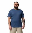 A bearded man with a tattoo on his left forearm stands facing forward in a blue Columbia Mens Zero Rules™ Light Technical T-Shirt and beige shorts, posing against a plain white background.