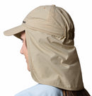 A person wears the Columbia Schooner Bank II Cachalot Hat in beige with a long neck flap for UPF 50 sun protection, seen from the side and slightly behind, set against a white background.