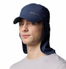 A person wearing a Columbia Schooner Bank II Cachalot Hat in navy blue with UPF 50 sun protection and neck flap, along with a light gray shirt, poses against a plain white background.