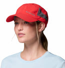 A woman in a Columbia Tech Shade II Cap, a red lightweight hiking hat with mesh side panels, and a light blue shirt looks slightly to the side against a white background.