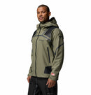 A man wearing a Columbia Mens Whistler Peak Waterproof Shell Jacket in olive green and black with black pants, standing and looking slightly to the side against a plain white background.