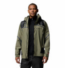 A man wearing a Columbia Mens Whistler Peak Waterproof Shell Jacket in olive green and black with zippered pockets, standing against a plain white background.