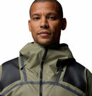 A man wears the Columbia Mens Whistler Peak Waterproof Shell Jacket in light olive-green with black shoulder patches and a high zippered collar, looking at the camera against a plain white background.