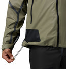 A hand pulls the adjustable drawcord at the hem of a Columbia Men’s Whistler Peak Waterproof Shell Jacket in olive green, highlighting the sleeve details and black pants.