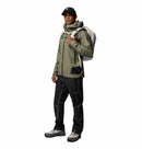 A man wearing a Columbia Mens Whistler Peak Waterproof Shell Jacket in green, black pants, hiking shoes, and a black cap stands facing forward with a beige backpack on his shoulder against a white background.