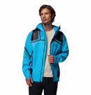 A man stands against a plain white background wearing a Columbia Mens Whistler Peak Waterproof Shell Jacket in bright blue with black accents, a white T-shirt underneath, and black pants.
