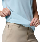A woman wearing beige pants and a Columbia Womens Zero Rules Light Technical T-shirt stretches the hem to display its sweat-activated cooling fabric. The photo highlights her torso and hands, with her face out of frame.