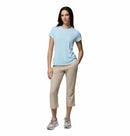 A woman stands against a white background wearing the Columbia Womens Zero Rules Light Technical T-shirt, featuring sweat-activated cooling. She pairs it with beige cropped pants and gray sneakers, one hand in her pocket and a slight smile on her face.
