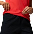 A person in a bright red Columbia Womens Zero Rules Light Technical T-shirt and black textured shorts lifts the hem with both hands, revealing sweat-activated cooling fabric detail.