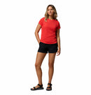 A woman with light brown hair poses against a white background in a Columbia Womens Zero Rules Light Technical T-shirt (red with sweat-activated cooling), black shorts, and black sandals—ideal for hiking. She has one hand in her pocket and one leg slightly bent.