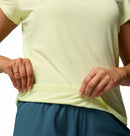A woman wears the Columbia Womens Zero Rules Light Technical T-shirt in light yellow and teal shorts, holding the hem to show its sweat-activated cooling feature—a perfect choice for hiking. Focus is on her torso and hands.