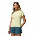 A woman stands against a white background wearing Columbia’s Womens Zero Rules Light Technical T-shirt in pale yellow and teal shorts, one hand in her pocket and smiling—featuring sweat-activated cooling, perfect for hiking adventures.