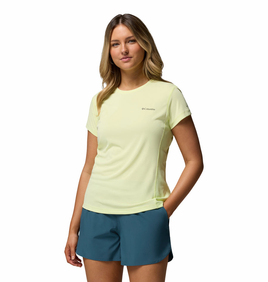 A woman stands against a white background wearing Columbia’s Womens Zero Rules Light Technical T-shirt in pale yellow and teal shorts, one hand in her pocket and smiling—featuring sweat-activated cooling, perfect for hiking adventures.