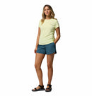 A woman with long, wavy blonde hair stands against a white background wearing the Columbia Womens Zero Rules Light Technical T-shirt, teal shorts, and black sandals—her left hand in her shorts pocket—perfect for hiking or staying cool.