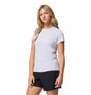 A woman with long blonde hair wears the Columbia Womens Zero Rules Light Technical T-shirt in light grey and black shorts, standing with one arm relaxed by her side against a plain white background—ideal for hiking or everyday wear.