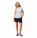 A woman with long, wavy blonde hair stands against a white background, wearing the Columbia Womens Zero Rules Light Technical T-shirt in light gray, paired with black shorts and gray athletic shoes, ready for outdoor adventures.
