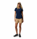 A woman with long blonde hair faces forward in a Columbia Womens Zero Rules Light Technical T-shirt (navy), beige shorts, beige cap, and black sneakers—ready for hiking against a plain white background.