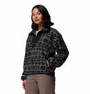 A woman with shoulder-length dark hair wears a Columbia Womens Sequoia Grove™ Printed Half Zip Fleece, featuring a black and white pattern, and light brown pants. She stands with hands in her pockets, looking to the side against a plain white background.