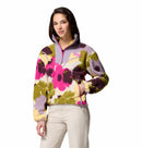 A woman wears the Columbia Womens Sequoia Grove™ Printed Half Zip Fleece, featuring a bold floral pattern and made from recycled polyester, paired with light beige pants as she stands with a hand in her pocket against a white background.