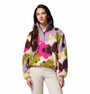 A young woman with long brown hair models the Columbia Womens Sequoia Grove™ Printed Half Zip Fleece in a bold pink, purple, yellow, and green floral print, paired with light pants against a white background.