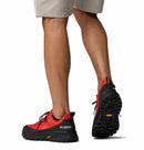 A person wearing tan shorts and red Columbia Mens Tellurix Titanium Outdry Hiking Shoes with black Vibram Megagrip soles stands on one leg, showing the rugged outsole of this waterproof hiking shoe from behind.