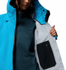 A person with face partially out of frame wears the Columbia Womens Saudan Pro™ 3L Shell Jacket in blue over a dark blue fleece, holding the shell open to reveal a zippered inner pocket. Features Omni-Tech Pro for extreme weather protection.