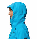 A woman in profile wears the Columbia Womens Saudan Pro™ 3L Shell Jacket, fully zipped with the hood up. The bright blue, seam-sealed shell has a hood pull cord and offers extreme weather protection against a white background.