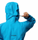 A woman adjusts the hood of a bright blue Columbia Womens Saudan Pro™ 3L Shell Jacket using the back drawstring; the visible Columbia logo highlights its advanced weather protection features.