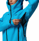A person in a bright blue Columbia Womens Saudan Pro™ 3L Shell Jacket unzips an underarm vent, revealing mesh lining. The shell features black zippers, a chest logo, and provides extreme weather protection.
